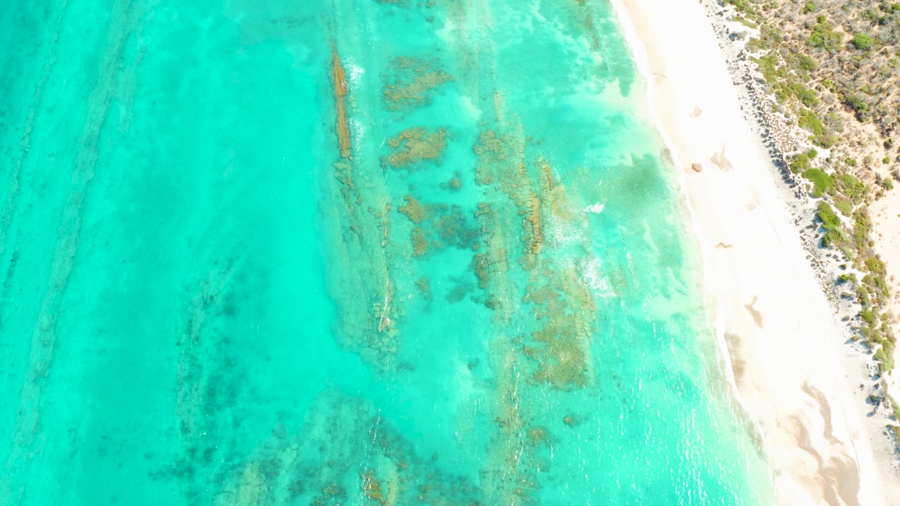 Aerial Cabo Pulmo frame showing shallow turquoise water, scattered submerged formations, pale sand on the right, scrubby land above the beach, and no visible people.