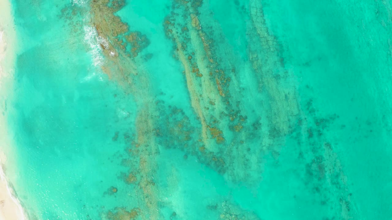 Overhead view of turquoise water in Cabo Pulmo with pale sand along the left edge, diagonal coral and seabed formations below the surface, and no visible people.