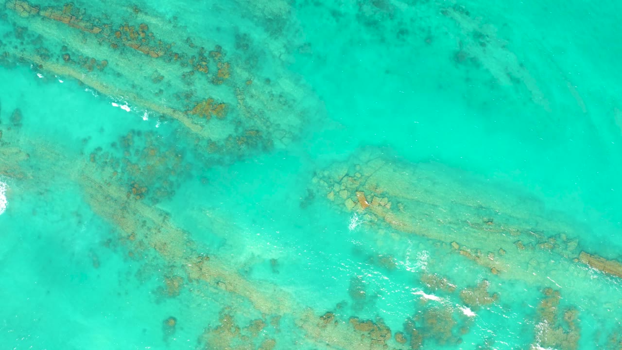 Aerial frame of Cabo Pulmo turquoise water with diagonal coral bands, bright ripple highlights near the lower center, and no visible people.