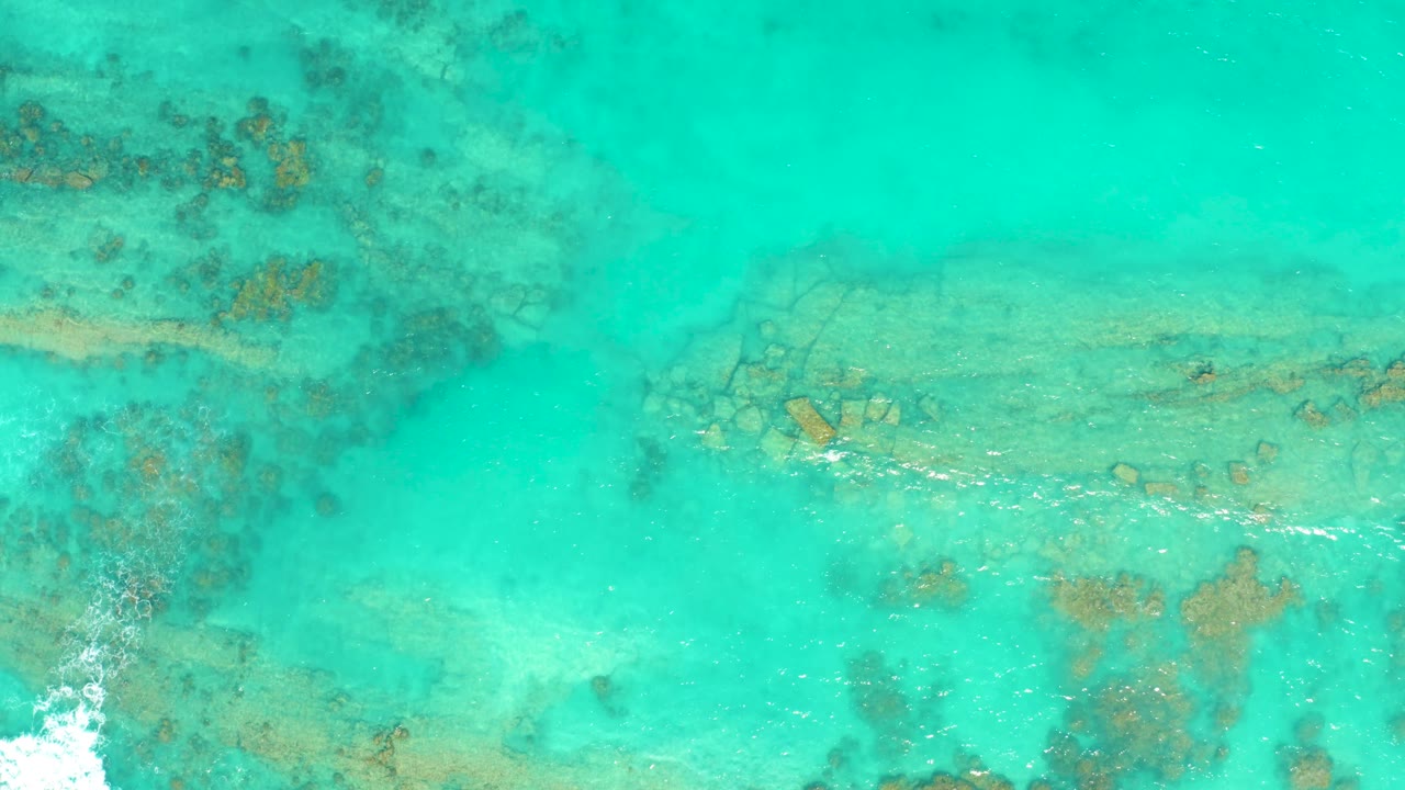 White foam enters near the lower left while clear Cabo Pulmo water reveals coral and rock formations across the frame, with no visible people.