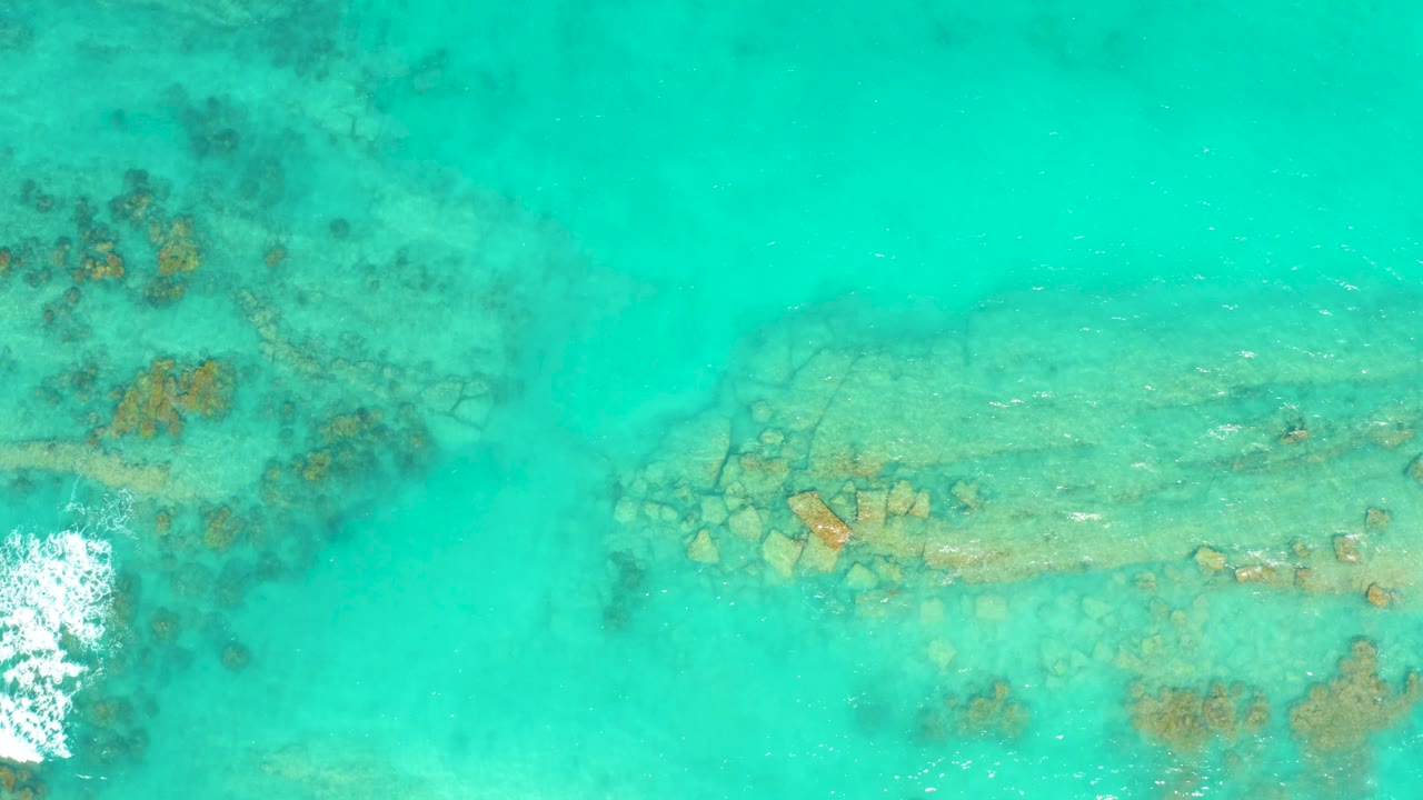 Overhead view of shallow turquoise water in Cabo Pulmo with reef patches beneath the surface, a pale seabed, white surf entering from the left edge, and no visible people.