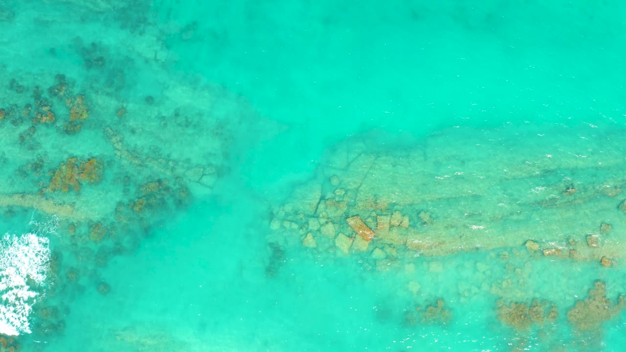 Overhead Cabo Pulmo frame showing blue-green shallows, submerged coral and rocks, a sandy bottom, and a bright white wave patch near the lower right, with no visible people.