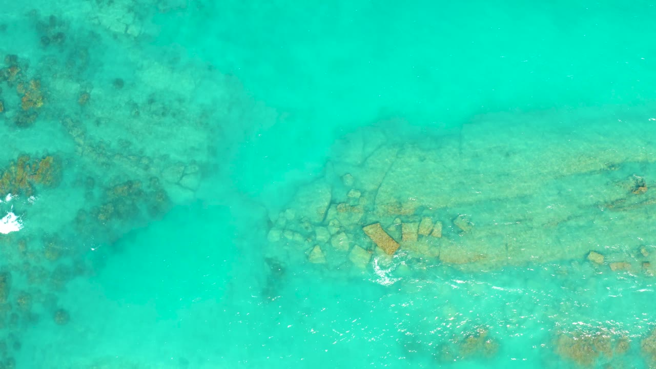 Overhead Cabo Pulmo water frame with a pale rock shelf below the surface and a brighter white splash forming near the lower center, no visible people.