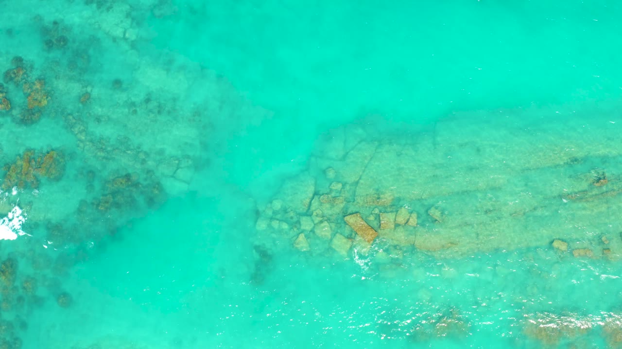 Top-down view of Cabo Pulmo's turquoise water with darker underwater patches, tan rocks below the surface, and a small white foam streak along the left side, no visible people.