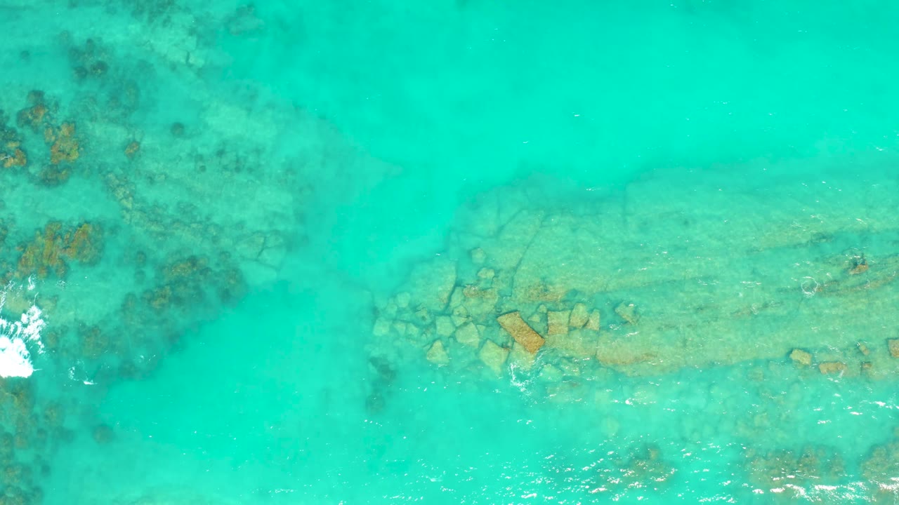 Cabo Pulmo shallow water seen from above with a long pale rock form under the surface and a white foam burst entering at the lower left, no visible people.