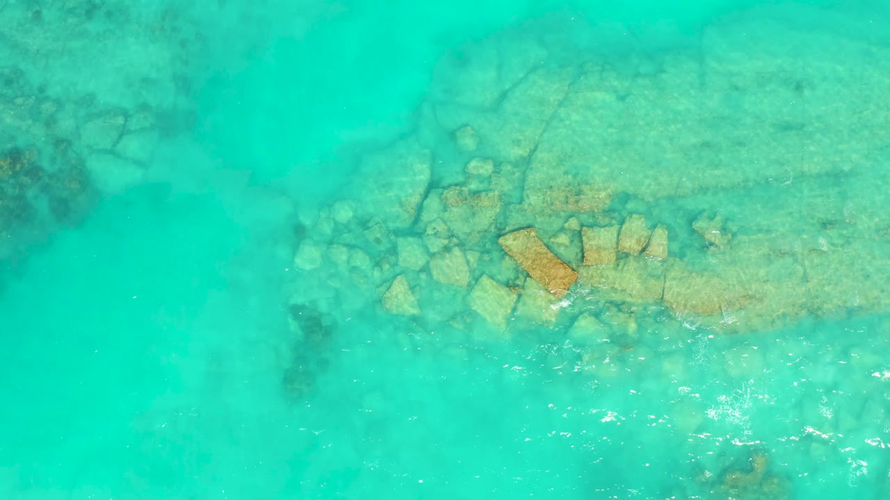 A top-down Cabo Pulmo water frame shows scattered underwater stones and a larger tan slab beneath turquoise ripples, with no visible people.