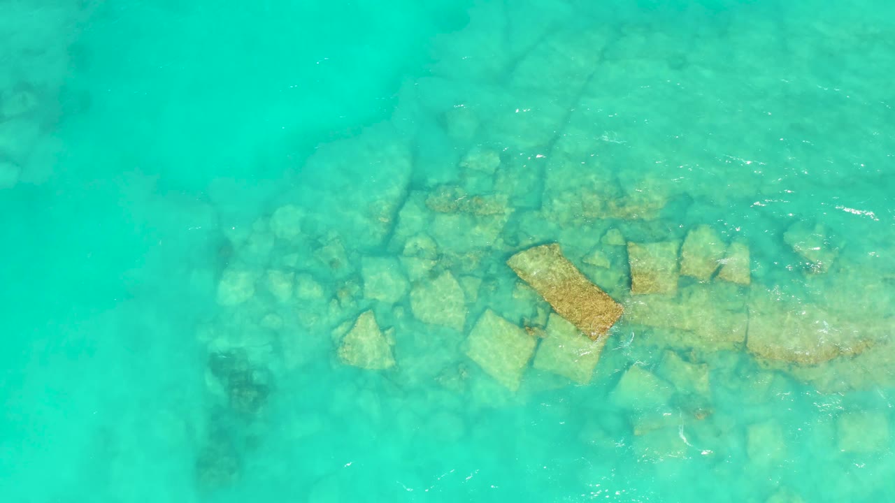Overhead Cabo Pulmo frame shows shallow turquoise ocean, submerged rocks, and a brighter ripple line crossing the right side, with no visible people.