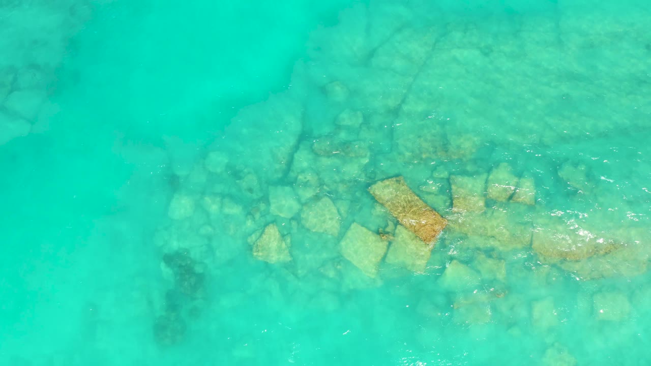 Cabo Pulmo water appears more green-blue in this overhead frame, with the central rock group still visible below the moving surface and no visible people.