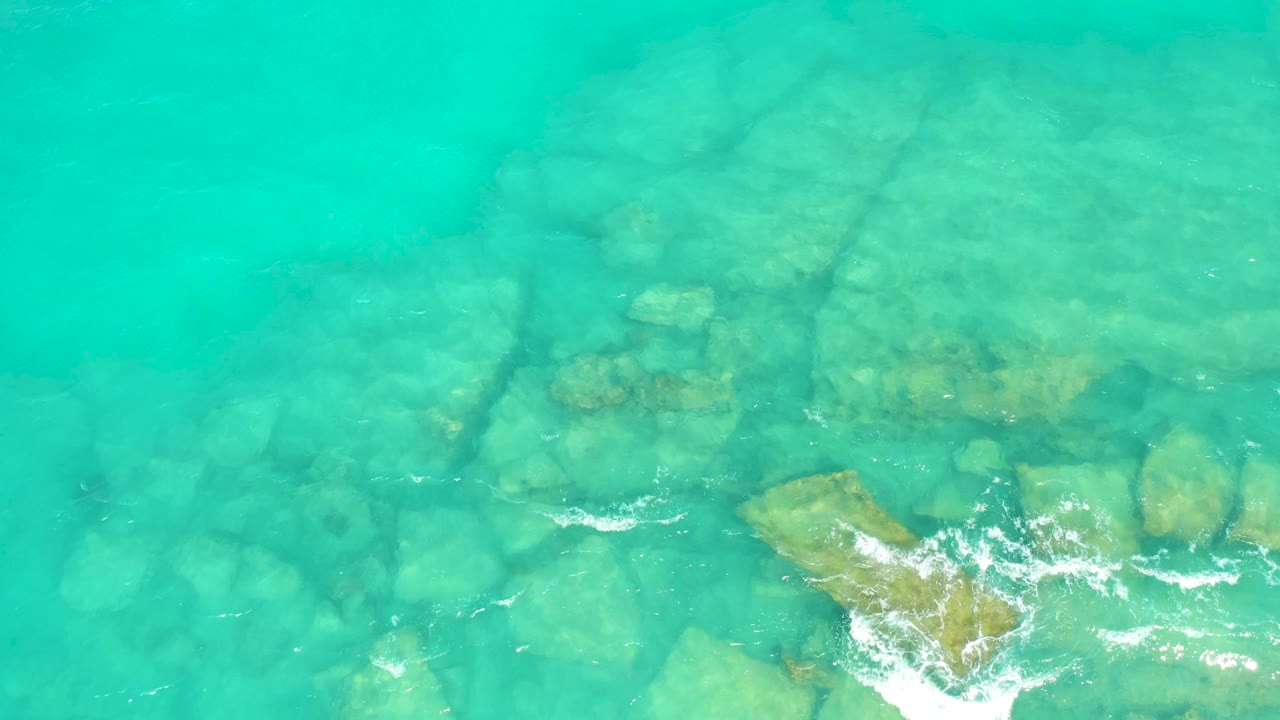 Overhead view of clear turquoise water in Cabo Pulmo, Mexico, with submerged rocks and a small patch of white foam near the lower edge, no visible people.