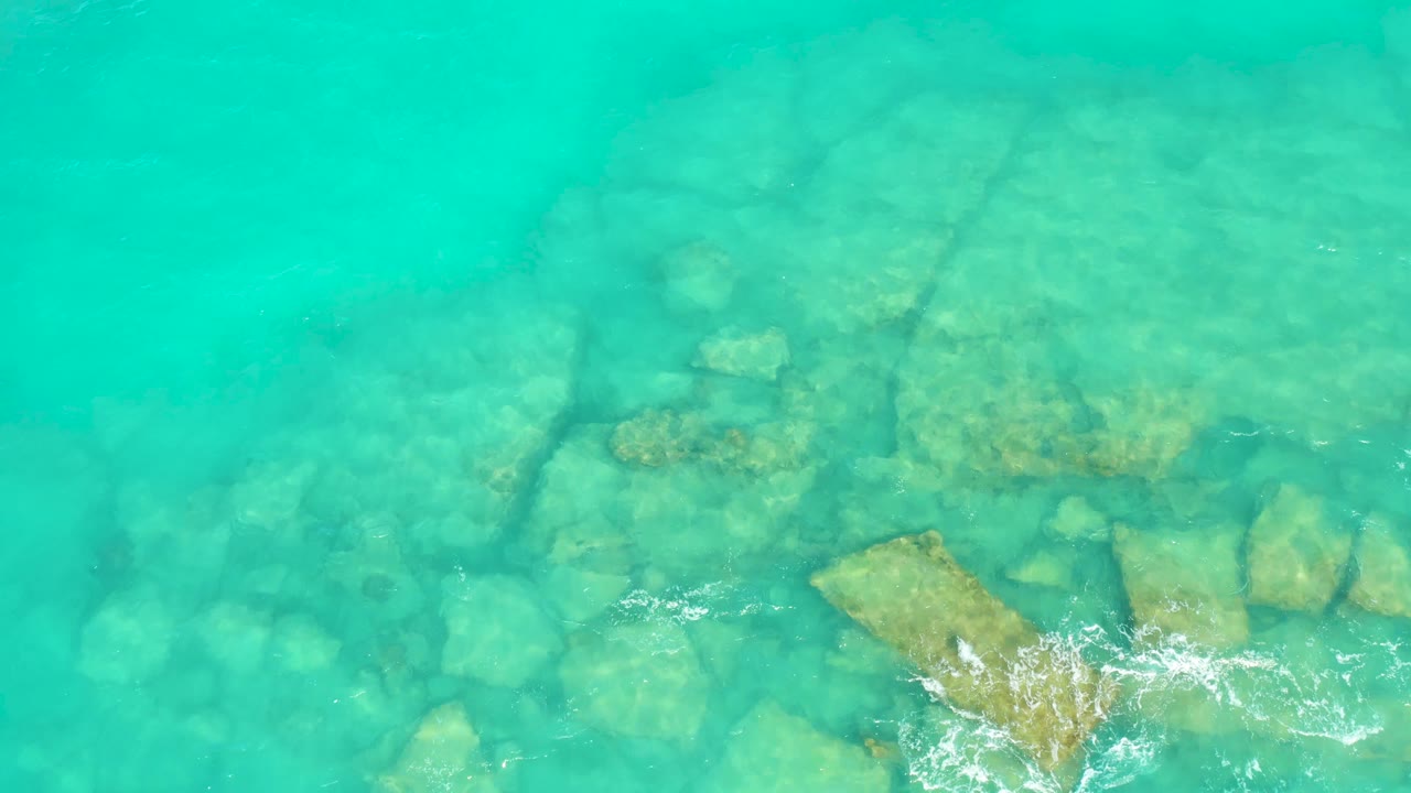 Clear overhead view in Cabo Pulmo with pale submerged rocks under turquoise water and white foam gathered closer to the lower right edge, no visible people.