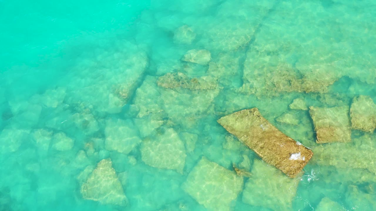 An overhead Cabo Pulmo frame shows the main tan stone block under turquoise water as a small white object or foam mark appears near its right edge, with no visible people.