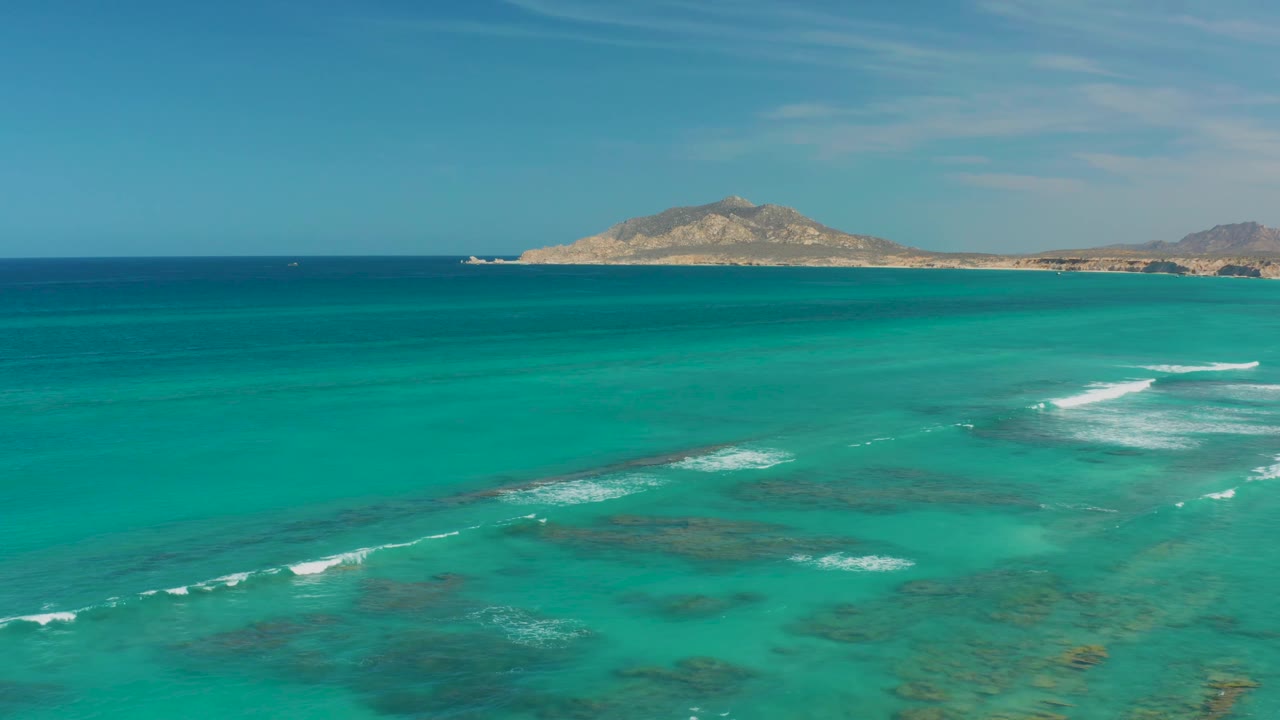 A wide Cabo Pulmo aerial view shows white waves spreading across shallow reef patches, deeper blue water beyond, dry island terrain on the horizon, and no visible people.
