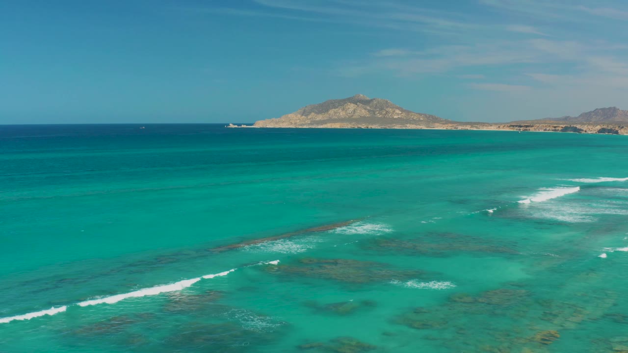 Mid-clip Cabo Pulmo frame with a stronger white surf line entering from the lower left, reef patches visible through turquoise water, distant island centered near the horizon, and no visible people.