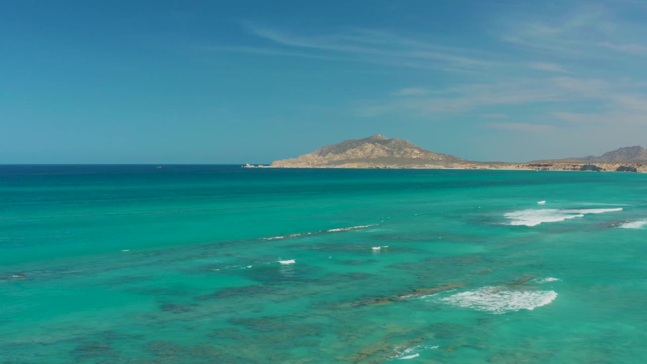 Wide Cabo Pulmo ocean frame showing turquoise shallows, a bright white patch of breaking surf to the right, distant rocky terrain, and no visible people.