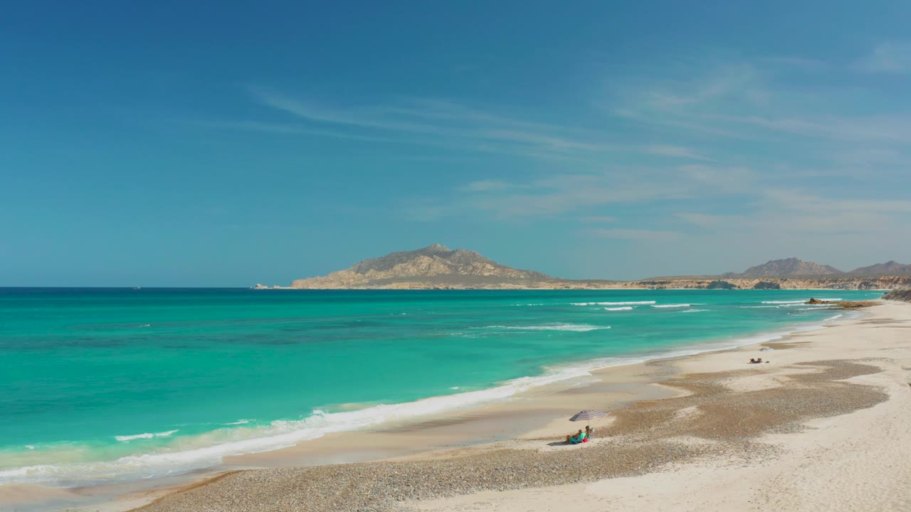 Cabo Pulmo beach view with turquoise surf, pale sand, a small umbrella and beachgoer near the lower right, and distant low hills beneath a clear blue sky.