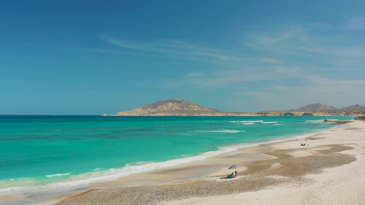 Wide turquoise shoreline with waves, sand, umbrella, and distant ridge.