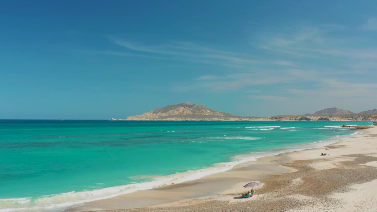 Cabo Pulmo beach with the umbrella and small beachgoer near the lower right edge, foamy waves moving over sand, and distant arid hills under blue sky.