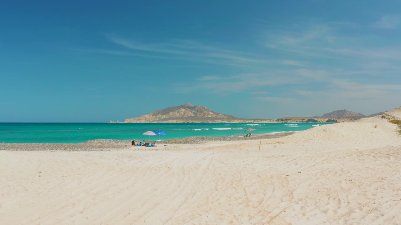 Wide Cabo Pulmo beach view with pale sand in the foreground, turquoise water beyond, a blue umbrella near distant beachgoers, and low mountains on the horizon.
