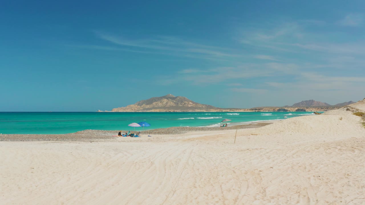 Wide Cabo Pulmo beach frame with the umbrella and beachgoers near the lower left, a clean turquoise band of ocean, light surf, pale dunes, and low mountains.