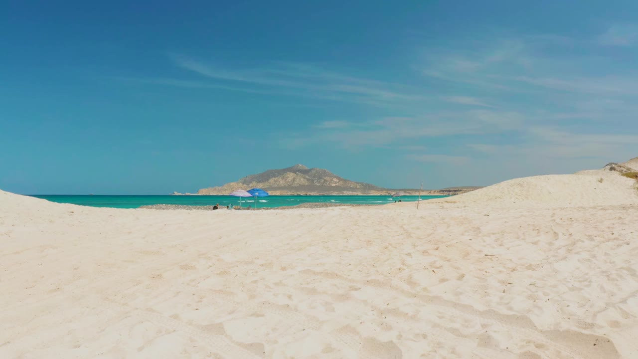 Wide Cabo Pulmo beach view at 0:00 with pale sand in front, turquoise ocean beyond, a blue umbrella near the shoreline, and distant hills under clear sky.
