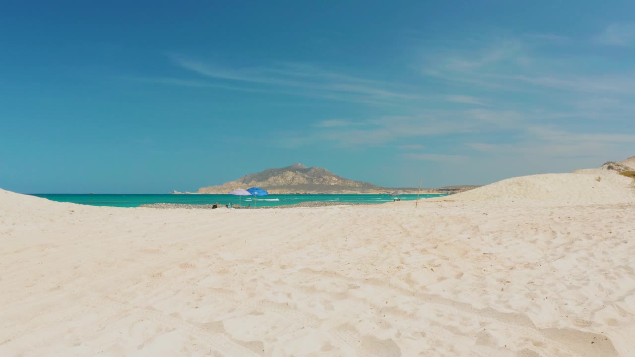 At 0:02, the Cabo Pulmo shoreline remains wide and quiet, with the blue umbrella near the water and low hills visible beyond the turquoise band.