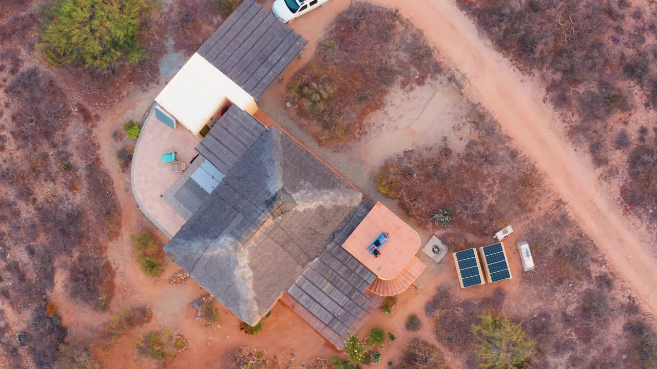 High overhead view of a thatched-roof house, patio areas, dirt road, and solar panels.
