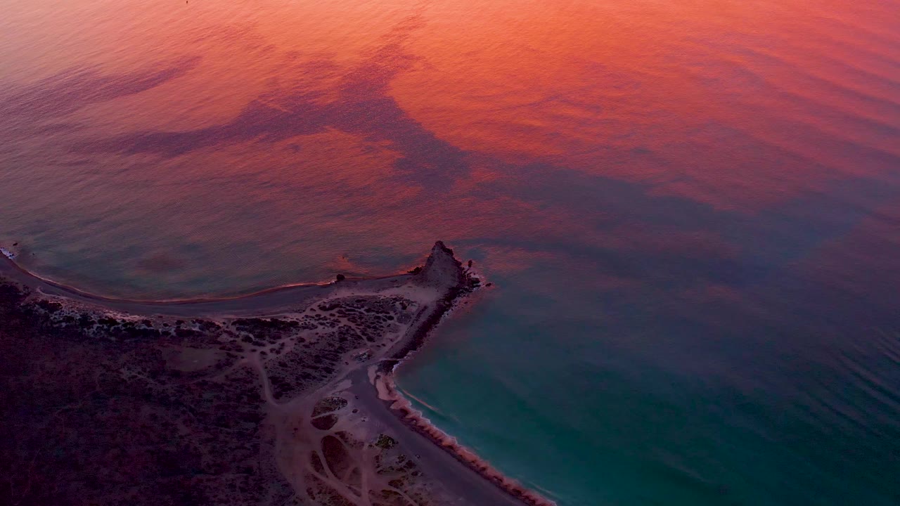 Aerial frame of the Cabo Pulmo lighthouse point with the shoreline curving around teal water while broad orange and pink reflection covers the upper ocean, with no visible people.