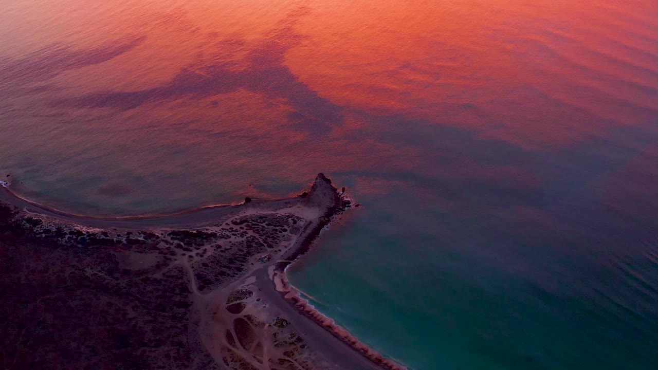 Cabo Pulmo aerial still with the lighthouse peninsula near the lower center, dark coastal terrain at left, turquoise water at right, and warm sunset color spreading across the upper sea, with no visible people.