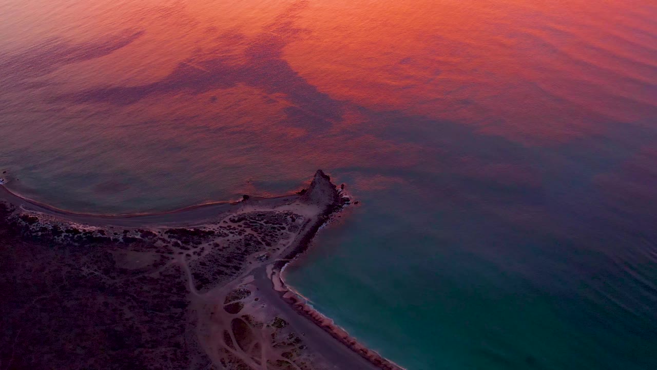 The lighthouse peninsula in Cabo Pulmo remains low in frame as darker land borders the shore and the ocean shows layered orange, pink, blue, and teal bands, with no visible people.
