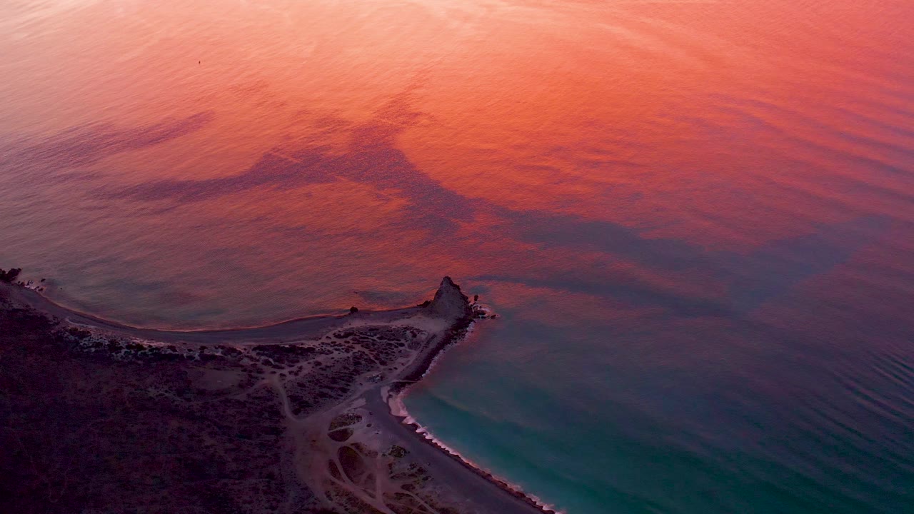 Aerial sunset view over Cabo Pulmo, Baja California Sur, with a dark coastal point below, turquoise water near shore, pink orange reflections across the sea, and no visible people.