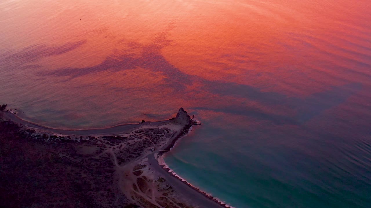 High aerial frame from Cabo Pulmo, Baja California Sur, with the breakwater angled into turquoise shallows while orange and pink sunset light spreads over the surrounding water, and no visible people.