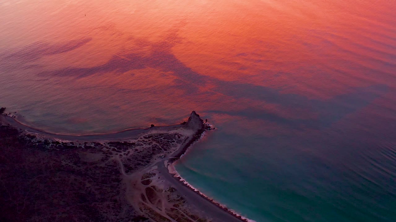 Aerial view of a dark coastal edge and breakwater below rippled sunset-colored water.