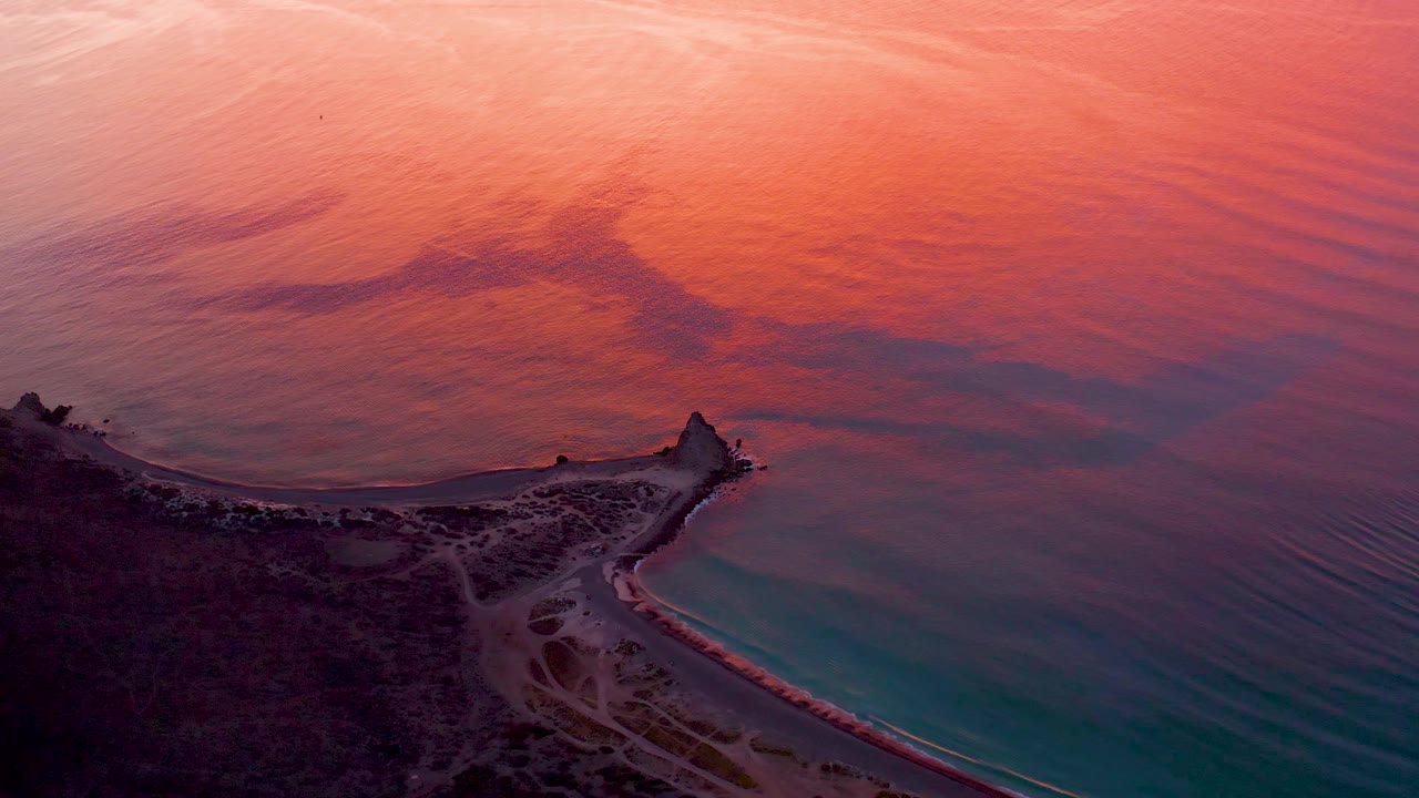 Aerial dusk frame over Cabo Pulmo showing a coastal point below broad pink-orange water reflection, turquoise water near the beach, faint shore paths, and no visible people.