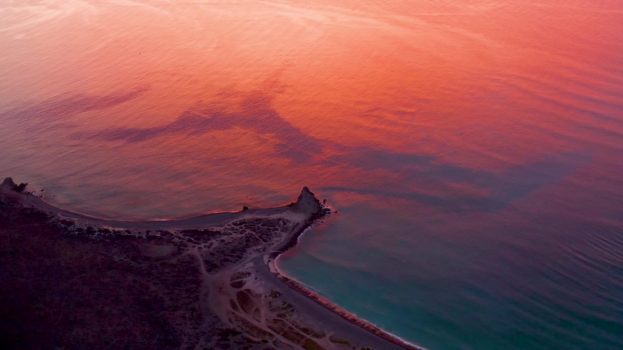 Aerial frame from Cabo Pulmo at dusk, with pink-orange water fading toward turquoise near the beach, visible shoreline paths on the land, and no visible people.