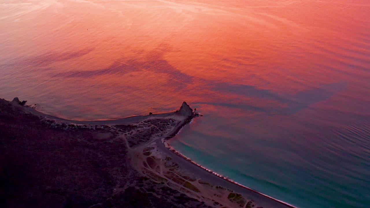 Aerial coastline view with a beach path, small vehicle, and warm reflected water.