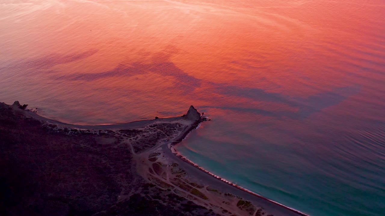 Cabo Pulmo aerial view showing dark land in the lower frame, a pale sandy track near the water, turquoise shallows along the shore, pink-orange ocean beyond, and no visible people.