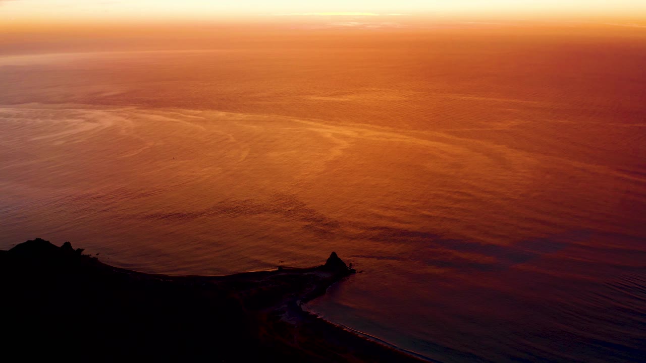 Cabo Pulmo sunset frame with broad orange water, faint wave lines, a high horizon, and black coastal silhouette along the bottom, no visible people.