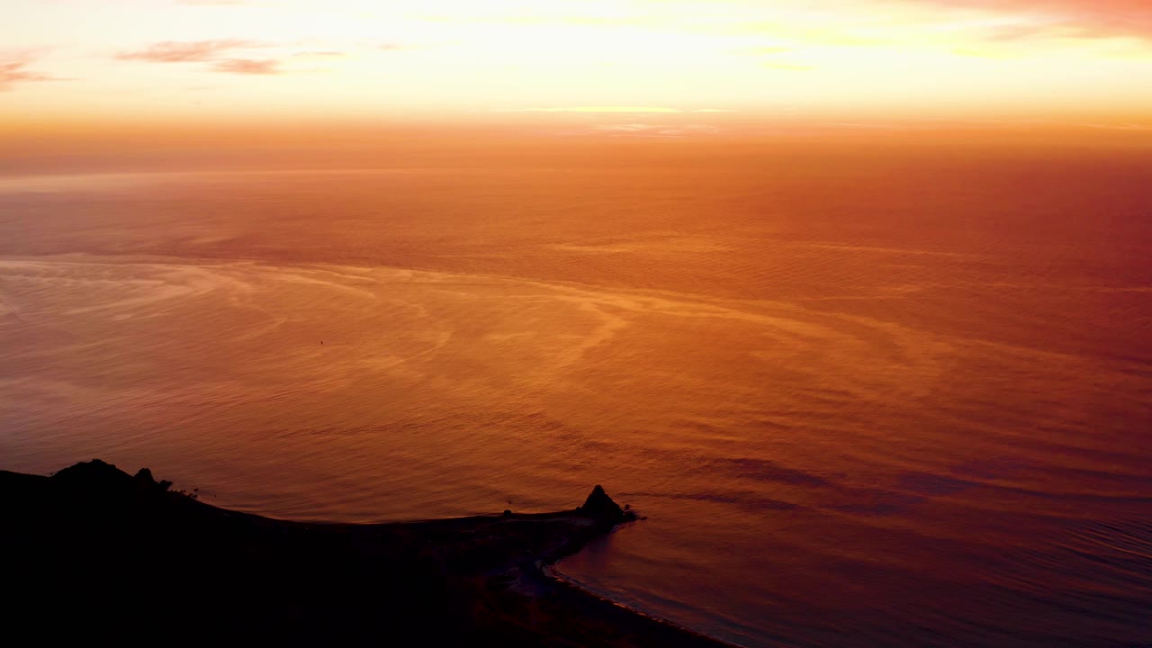Cabo Pulmo ocean sunset frame with broad orange reflections on rippled water, a dark foreground headland, pale yellow sky near the horizon, and no visible people.