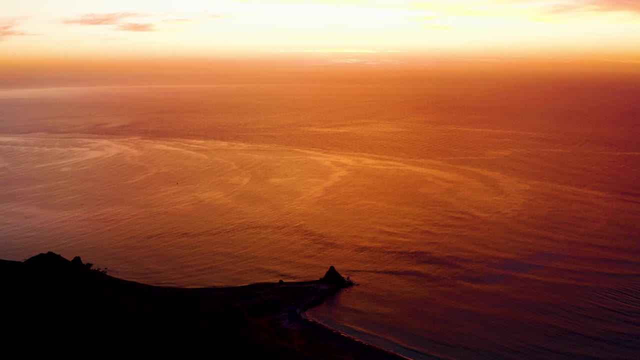 Cabo Pulmo evening frame showing a dark shore and cliff at the lower edge, warm orange ocean texture, soft yellow horizon light, and no visible people.