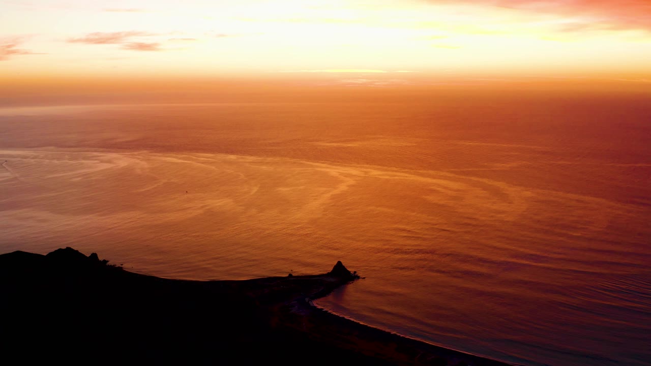 Cabo Pulmo sunset view with orange ocean reflections, a dark shoreline and small lighthouse silhouette at the lower edge, and no visible people.