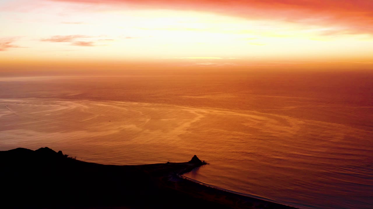 Orange sunset over calm ocean with dark coastal silhouette.