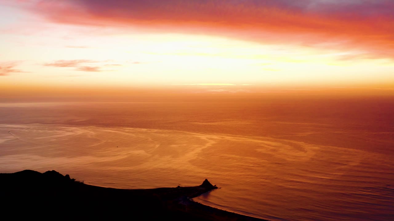 Cabo Pulmo sunset midway through the clip, showing orange water patterns, a pale glowing horizon, and the dark cliff edge running across the lower foreground with no visible people.