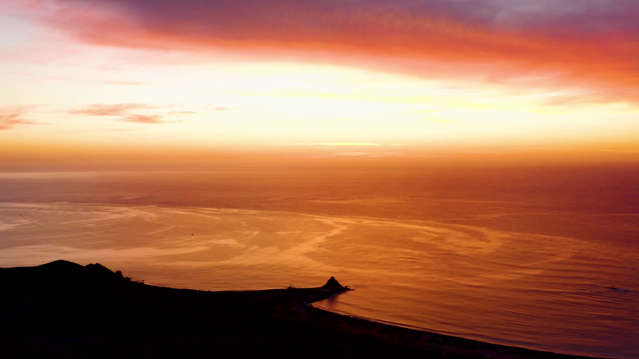 Cabo Pulmo ocean horizon glowing orange and yellow, with rippled reflections on the water and dark land cutting across the bottom edge, no visible people.