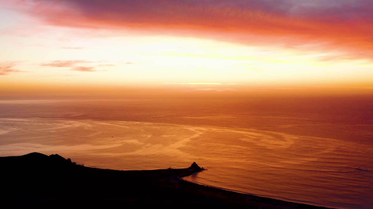 Cabo Pulmo sunset frame with a broad pink cloud band above amber ocean water and a black cliff shape along the lower frame, no visible people.