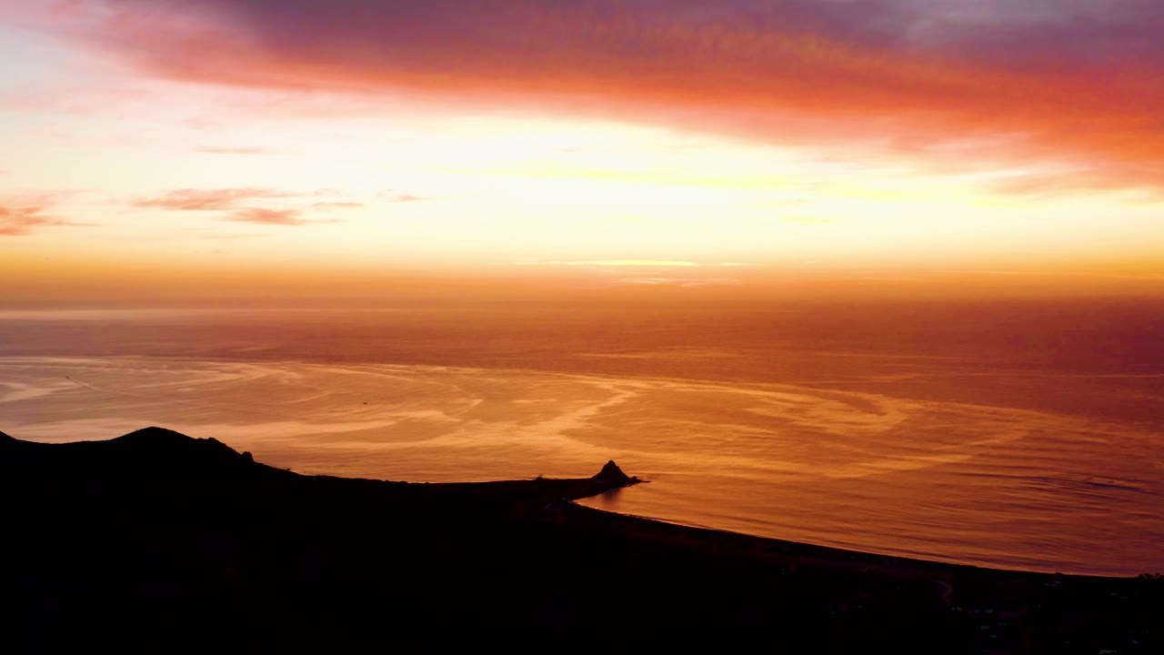 At 0:02, layered pink clouds sit over a gold-orange ocean surface and the Cabo Pulmo shoreline stays in black silhouette, with no visible people.