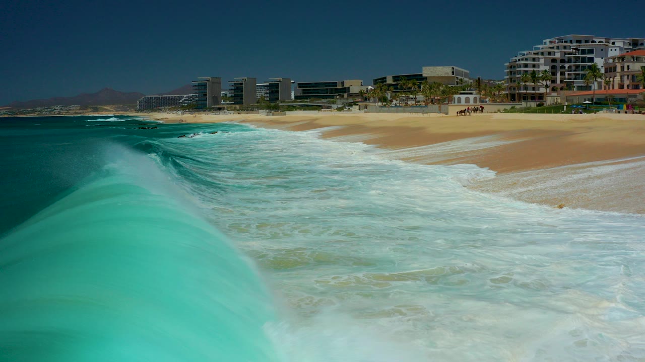 At Marquis Los Cabos, a turquoise wave rises in the foreground near a sandy beach, with coastal buildings and clear sky in the distance and no discernible people.