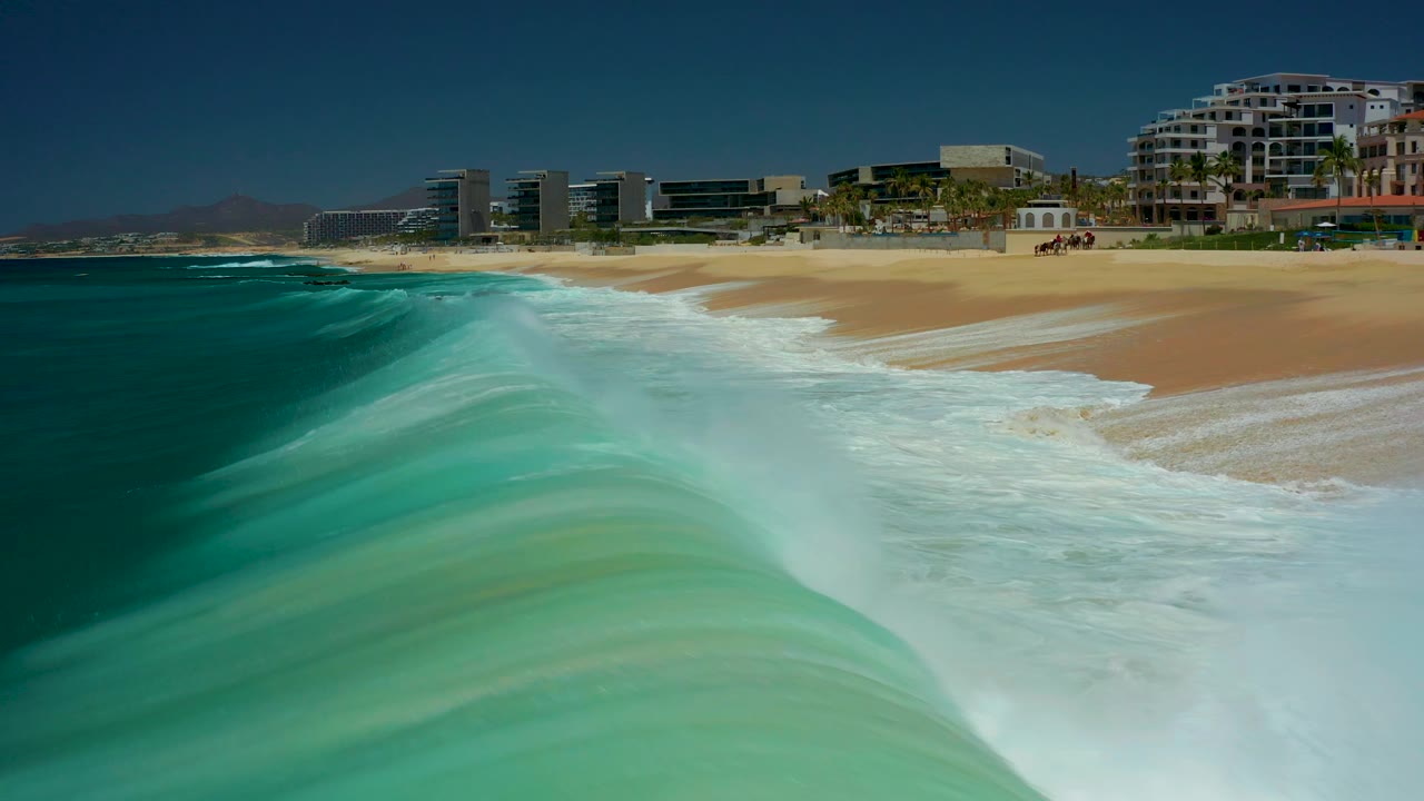 At Marquis Los Cabos, the wave face dominates the foreground as white surf pushes along the beach, while modern coastal buildings line the horizon and no discernible people are visible.