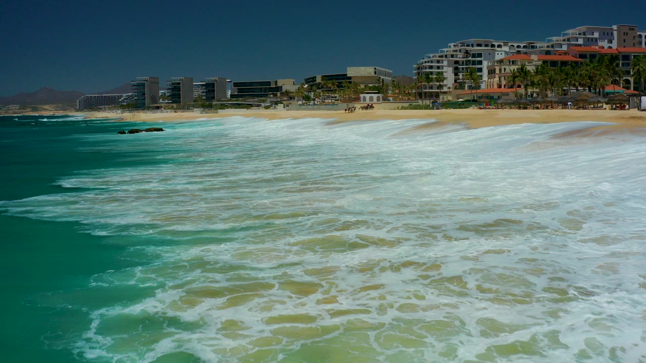 Turquoise surf and white foam rolling toward a sandy beach with coastal buildings beyond.