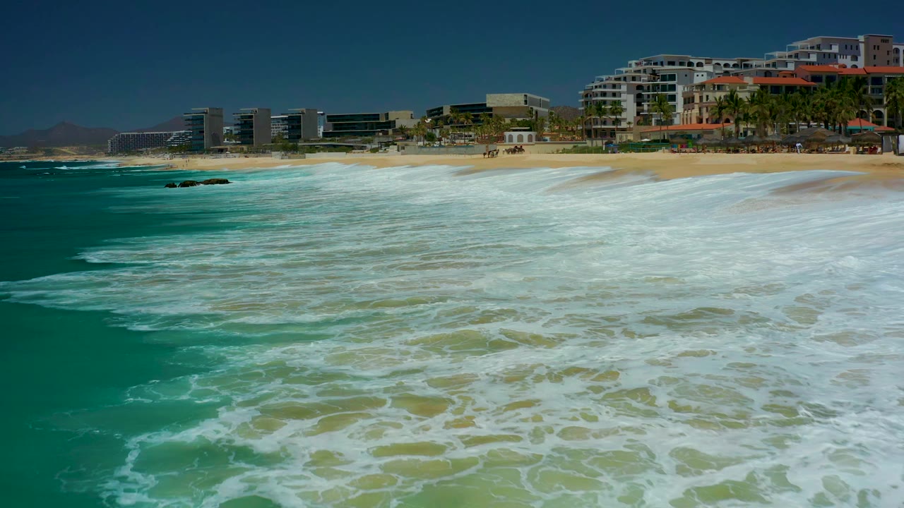At 0:01, the foamy turquoise surf sits slightly farther across the foreground, with tan beach and modern coastal buildings visible along the upper edge at Marquis Los Cabos.