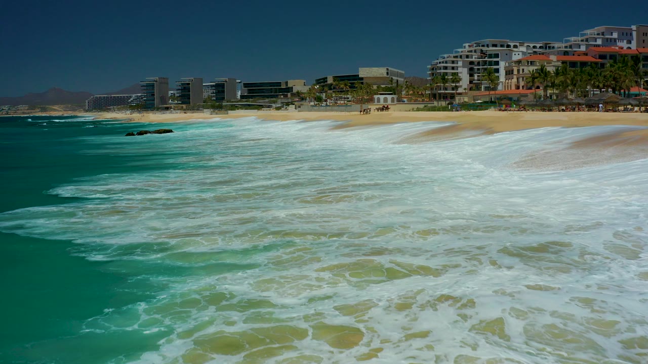 At 0:02, the wave foam forms a wide bright band across the lower frame while the sandy beach and resort buildings remain fixed in the background at Marquis Los Cabos.